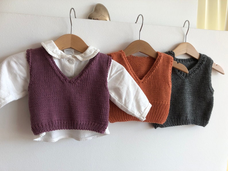 May include: Three knitted vests for children, one purple, one orange, and one grey. The vests are hanging on wooden hangers.