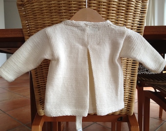 May include: White knitted baby sweater with a long back opening. The sweater is lying on a wicker chair.