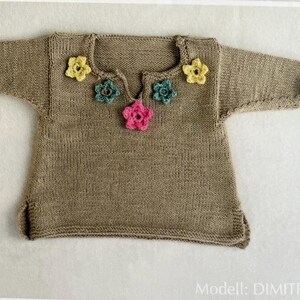 May include: A tan-colored, knitted top with short sleeves and a v-neckline. The neckline is adorned with five colorful, crocheted flowers in yellow, teal, and pink. The text "Modell: DIMITRA" is visible at the bottom.