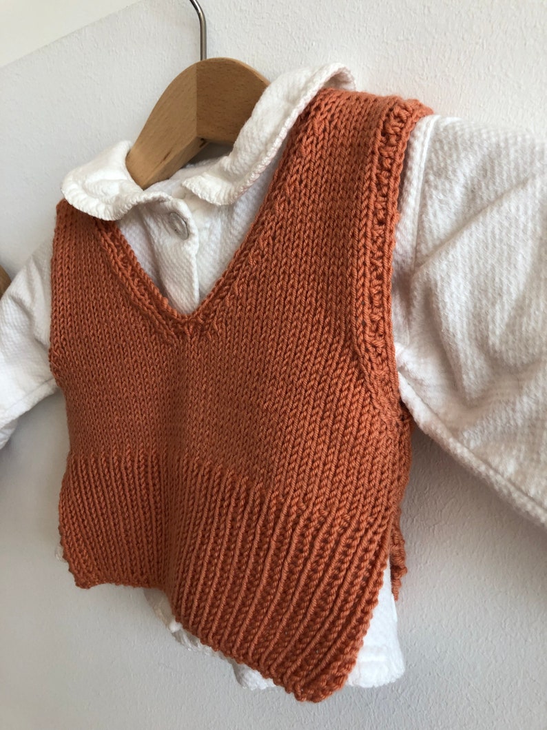 May include: A rust-coloured knitted waistcoat with a V-neckline. The waistcoat is hanging on a wooden hanger and is layered over a white collared shirt.