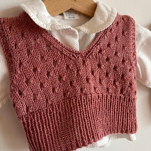 Pode incluir: Um colete de malha rosa com um padrão de renda delicado. O colete tem um decote em V e uma bainha canelada. O colete está pendurado em um cabide de madeira.