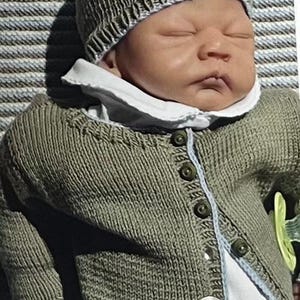 May include: A baby doll wearing a knitted olive green hat and cardigan with button closures. The doll has a white collared shirt underneath and a pacifier. The text "Modellserie: JOHANNES" is visible at the bottom.