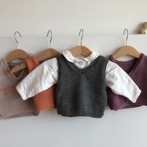 May include: Four knitted vests for children, hanging on hangers. The vests are in different colours: beige with orange lettering, orange, grey, and purple.