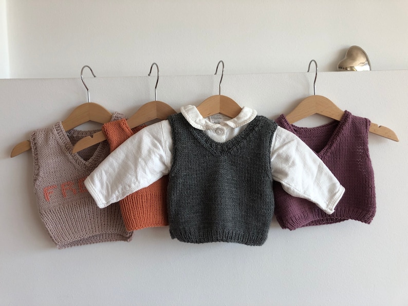 May include: Four knitted vests for children, hanging on hangers. The vests are in different colours: beige with orange lettering, orange, grey, and purple.