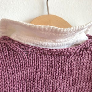 May include: A close-up of a mauve knitted jumper with a white ruffled collar. The jumper is hanging on a wooden hanger.