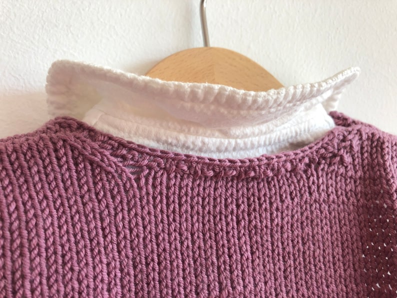 May include: A close-up of a mauve knitted jumper with a white ruffled collar. The jumper is hanging on a wooden hanger.