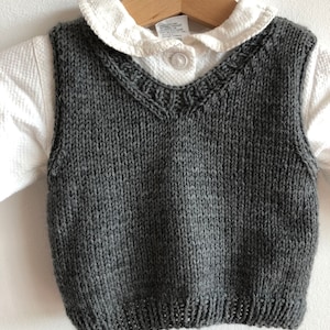 Könnte beinhalten: Eine graue Strickweste mit V-Ausschnitt und einem dekorativen Besatz am Halsausschnitt. Die Weste hängt an einem Holzbügel.