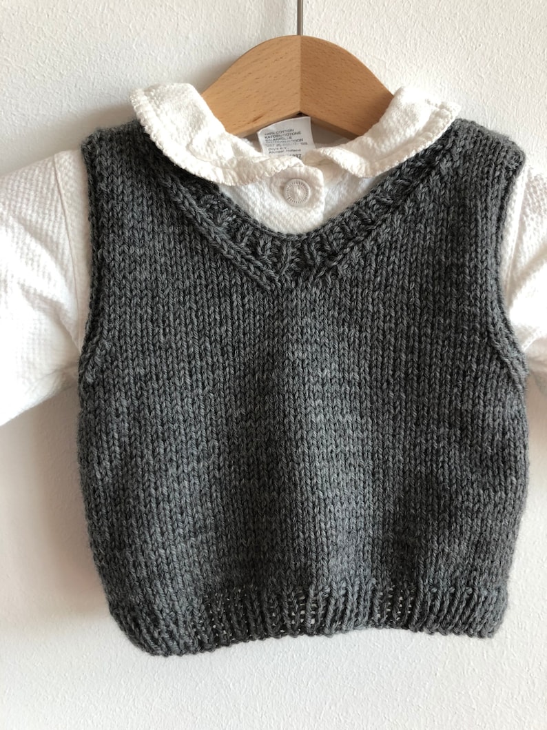 May include: A grey knitted waistcoat with a V-neck and a decorative trim around the neckline. The waistcoat is hanging on a wooden hanger.