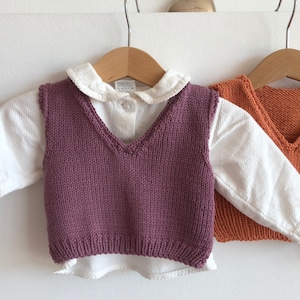 May include: Two knitted vests for children, one in purple and one in orange, hanging on hangers. Both vests have a V-neck and are made of a soft, textured yarn.