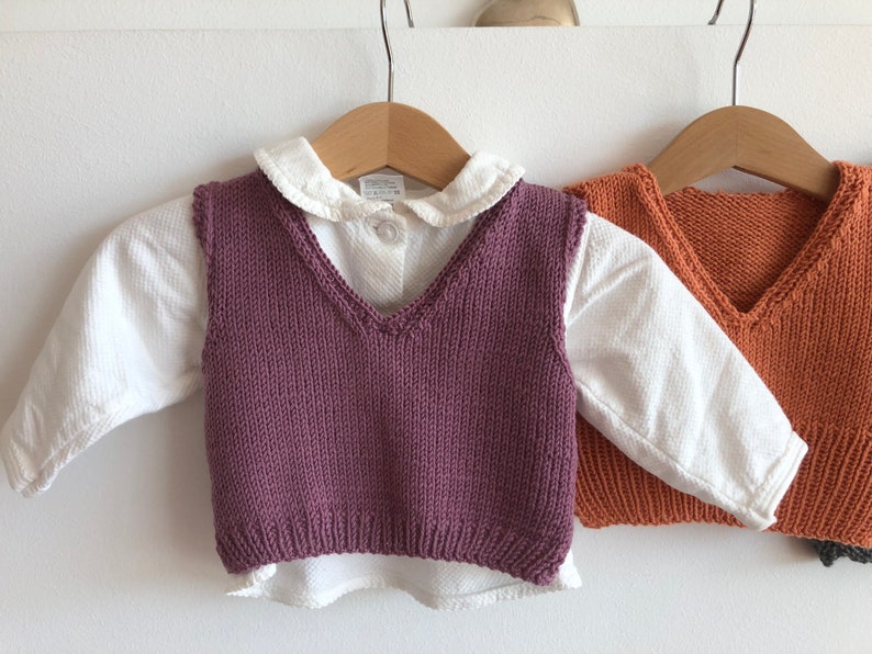 May include: Two knitted vests for children, one in purple and one in orange, hanging on hangers. Both vests have a V-neck and are made of a soft, textured yarn.