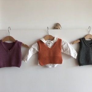 May include: Three knitted vests hanging on hangers. The vest on the left is purple, the middle vest is orange, and the vest on the right is grey.