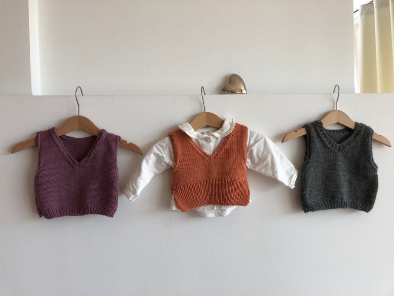 May include: Three knitted vests hanging on hangers. The vest on the left is purple, the middle vest is orange, and the vest on the right is grey.