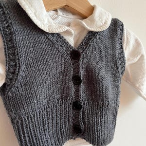 Könnte beinhalten: Eine graue Strickweste mit einem weißen Hemd mit Kragen darunter. Die Weste hat einen V-Ausschnitt und Knöpfe auf der Vorderseite.
