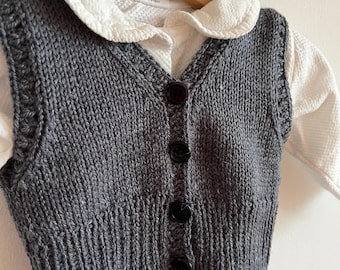 May include: A grey knitted waistcoat with a white collared shirt underneath. The waistcoat has a V-neck and buttons down the front.