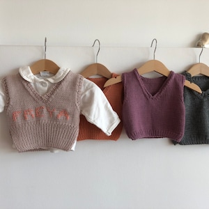 May include: Four knitted vests for children, hanging on hangers. One vest is beige with the name "FREYA" embroidered on the front in orange. Another vest is orange, one is purple, and the last one is grey.