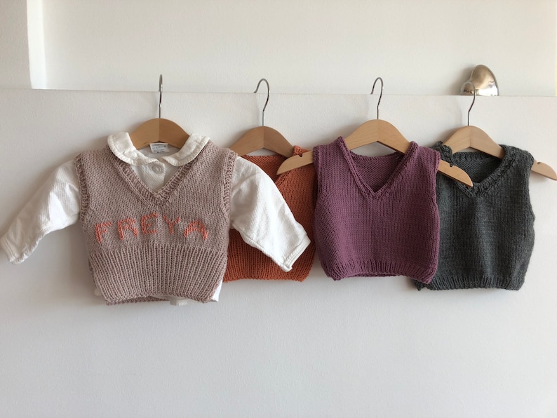 May include: Four knitted vests for children, hanging on hangers. One vest is beige with the name "FREYA" embroidered on the front in orange. Another vest is orange, one is purple, and the last one is grey.