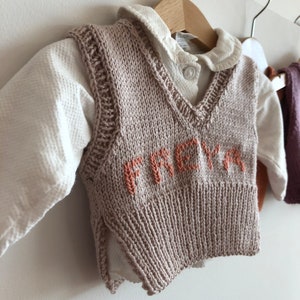 May include: A beige knitted waistcoat with a V-neck and the name "FREYA" embroidered in orange on the front.