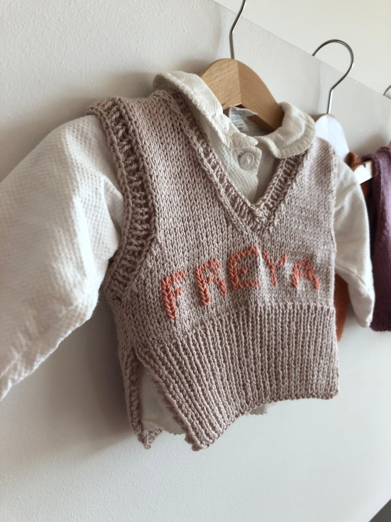 May include: A beige knitted waistcoat with a V-neck and the name "FREYA" embroidered in orange on the front.