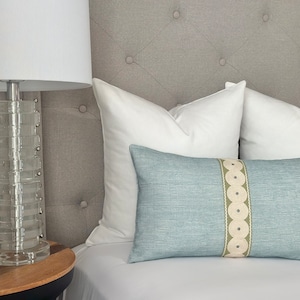 May include: A light blue throw pillow with a white and green patterned stripe. The pillow is on a bed with white bedding and a gray headboard.