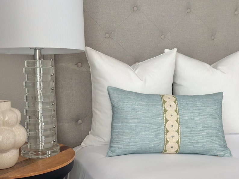 May include: A light blue throw pillow with a white and green patterned stripe. The pillow is on a bed with white bedding and a gray headboard.