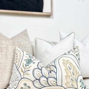 May include: Cushions with floral and paisley designs in blue, green, and beige. The cushions are on a white surface, with other cushions in the background. These cushions add a touch of elegance to any living space.