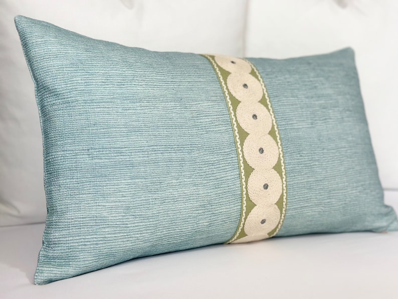 May include: A light blue throw pillow with a decorative green and white trim. The trim features a repeating pattern of circles.