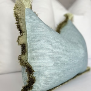 May include: A light blue pillow with a green fringe border. The pillow is lying on a white surface.