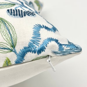以下が含まれることがあります： 青と緑の刺繍の花柄が施された白い枕カバー。枕カバーはジッパーで閉じられます。