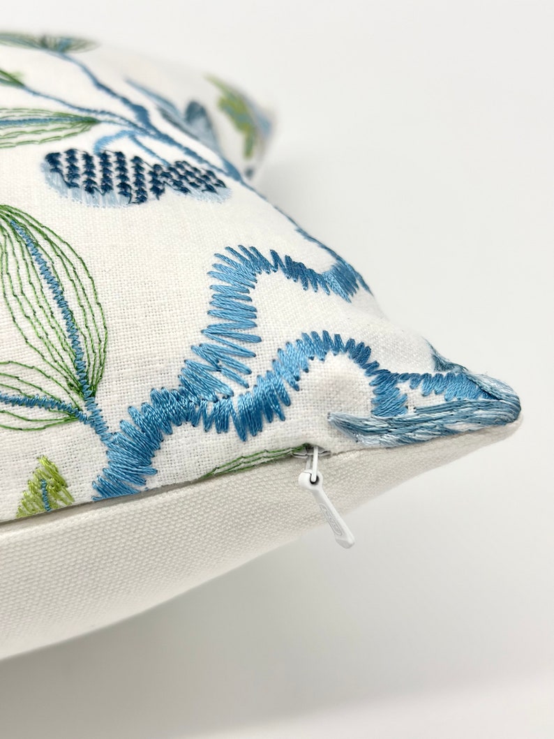 以下が含まれることがあります： 青と緑の刺繍の花柄が施された白い枕カバー。枕カバーはジッパーで閉じられます。