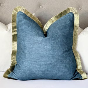 May include: A blue throw pillow with a green fringe border. The pillow is sitting on a white bed.