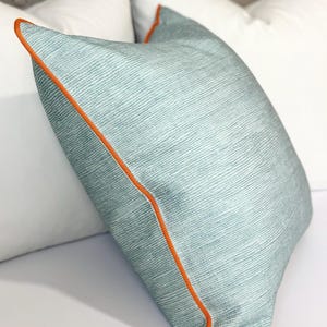 Puede incluir: Una almohada de color azul claro con un patrón texturizado sutil y un borde naranja.