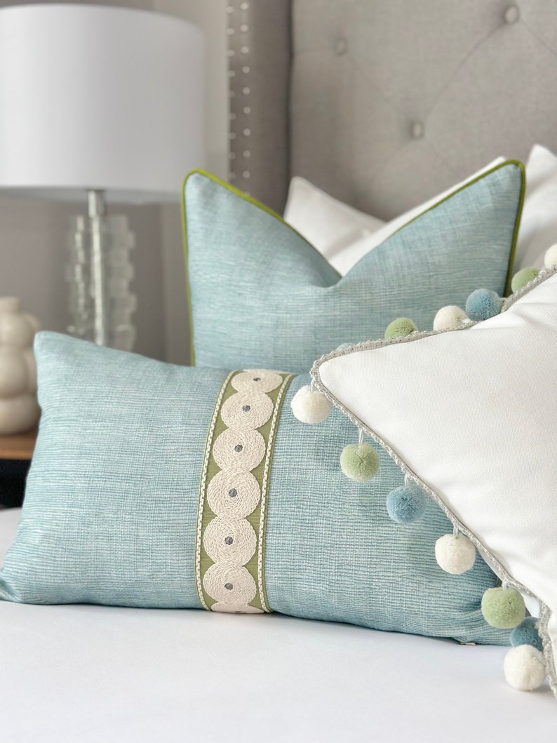 May include: A light blue throw pillow with a white and green trim and a decorative white ribbon with a circle pattern. The pillow is on a bed with a white bedspread and a light blue and green pillow behind it.
