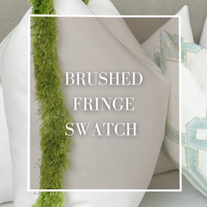 May include: A white pillow with a green brushed fringe trim. The pillow is on a light gray tufted headboard. The text "BRUSHED FRINGE SWATCH" is printed on the pillow.