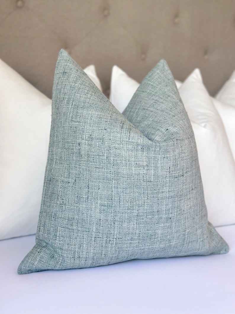 K&ouml;nnte beinhalten: Ein hellblaues Kissen mit strukturiertem Stoff. Das Kissen liegt auf einem wei&szlig;en Bett mit einem hellbraunen Kopfteil.