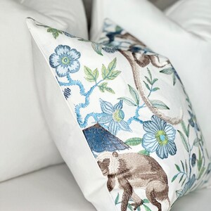 以下が含まれることがあります： 青と緑の花柄が刺繍された白い枕。茶色の猿が刺繍されています。