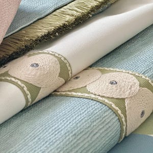May include: Close-up of fabric swatches with a light blue, white, and green color scheme. The fabric has a textured surface and a decorative trim with embroidered circles.