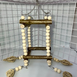 May include: A wooden bird toy with a square frame and a chain for hanging. The frame is decorated with white wooden beads. The toy has three hanging woven rope toys.