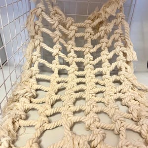 May include: Cream-coloured macrame hammock with a woven net design. The hammock is made of thick, twisted rope and features knotted intersections. The edges are finished with fringe. The hammock is displayed inside a white wire basket.