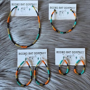 May include: A set of beaded jewelry featuring a necklace and earrings. The necklace and earrings are made with black, orange, gold, and turquoise beads. The jewelry is displayed on white cards with the text "RODEO RAT COMPANY" and the hashtag "#rodeoratco".