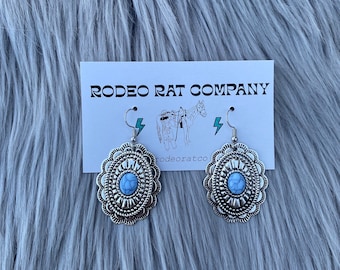 Puede incluir: Pendientes de plata con una piedra turquesa y un diseño decorativo. Los pendientes están sobre un fondo de piel sintética blanca. El texto "RODEO RAT COMPANY" está en una tarjeta blanca detrás de los pendientes.