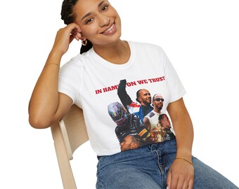 May include: A white t-shirt featuring a graphic print of a group of people, including someone wearing a racing helmet. The text "IN HAMILTON WE TRUST" is printed on the shirt.
