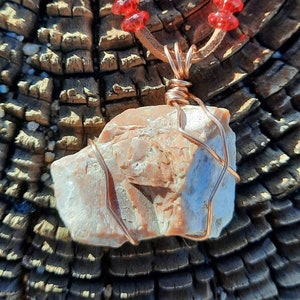 May include: A necklace with a light pink and white stone pendant wrapped in copper wire. The necklace is made with brown leather cord and red glass beads.