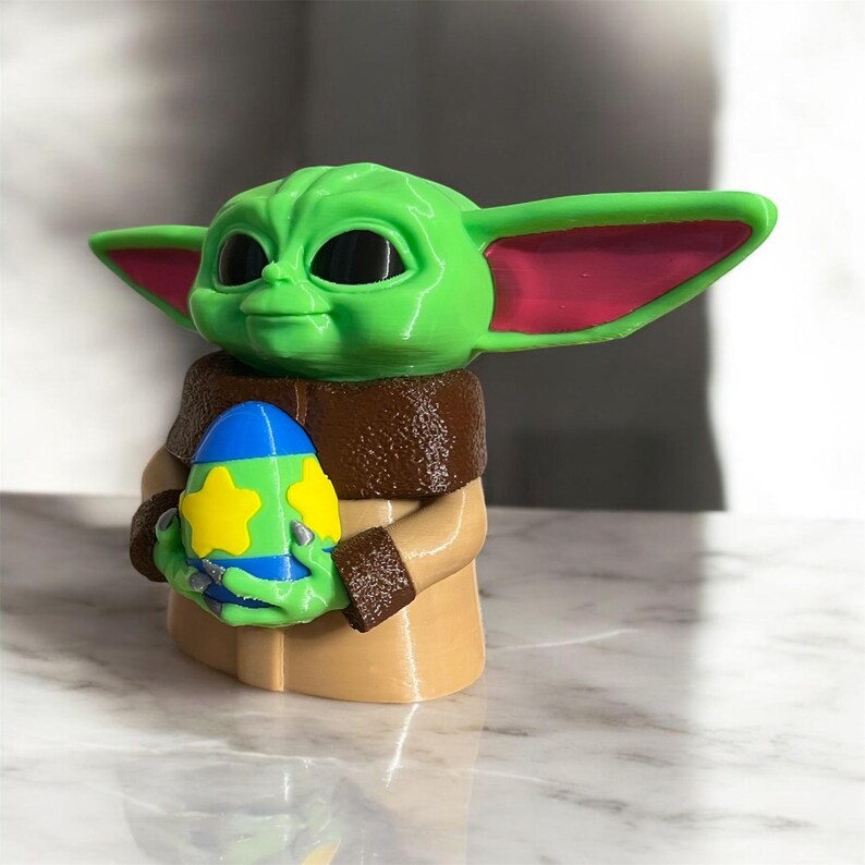 May include: A green and brown 3D printed figurine of a character from the Star Wars franchise holding a blue, green, and yellow Easter egg.