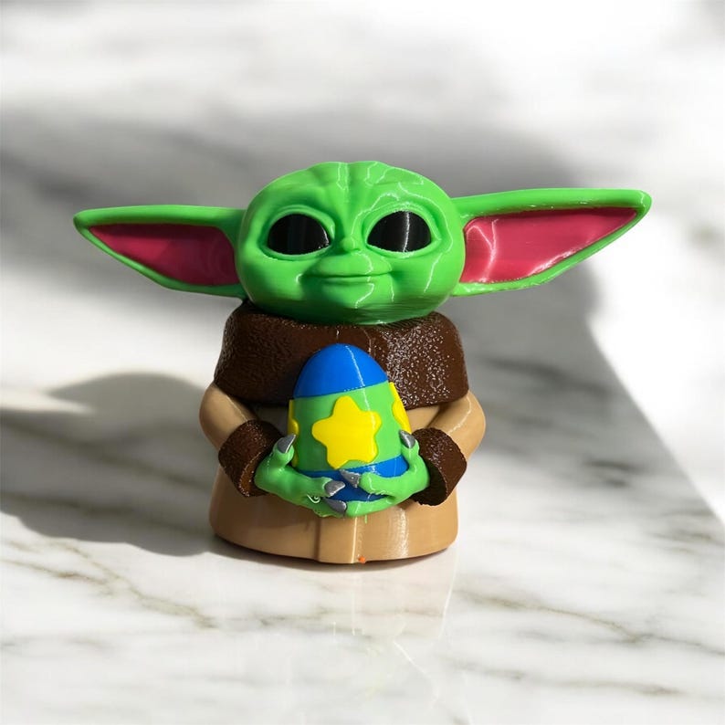 May include: A green and pink figurine of a character from the Star Wars franchise, holding a blue, yellow, and green egg-shaped object.