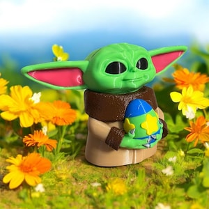 May include: A green and pink figurine of a character from the Star Wars franchise holding a blue and yellow Easter egg. The figurine is sitting in a bed of green grass and yellow flowers.