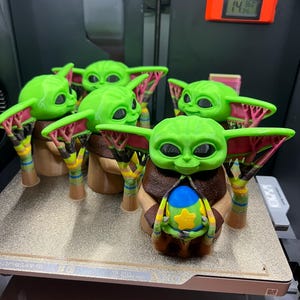 May include: Five green 3D printed figurines of a popular character from the Star Wars franchise. The figurines are holding a small blue and yellow object. The figurines are on a white surface with a black and silver background.