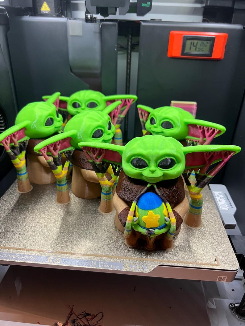 May include: Five green 3D printed figurines of a popular character from the Star Wars franchise. The figurines are holding a small blue and yellow object. The figurines are on a white surface with a black and silver background.