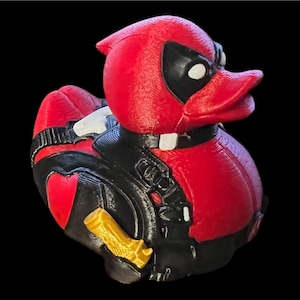 May include: A red and black rubber duck figure designed to resemble a costumed character. The duck features a black mask around the eyes, a black utility harness with white accents, and a small gold-colored weapon attached to its side.