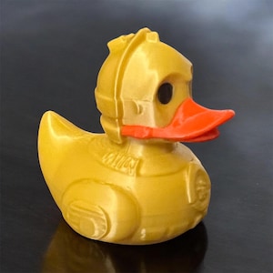 Könnte beinhalten: Eine gelbe Quietscheente mit orangefarbenem Schnabel und schwarzen Augen. Die Ente trägt einen goldfarbenen Helm und eine Rüstung, was ihr ein roboterhaftes Aussehen verleiht. Ein originelles Spielzeug.