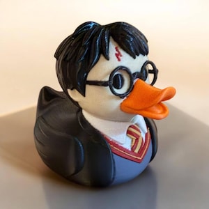 May include: A rubber duck designed to resemble a character with black hair, a red lightning bolt scar on the forehead, and round black glasses. The duck has an orange beak and wears a black robe over a white shirt with a red and yellow striped tie.